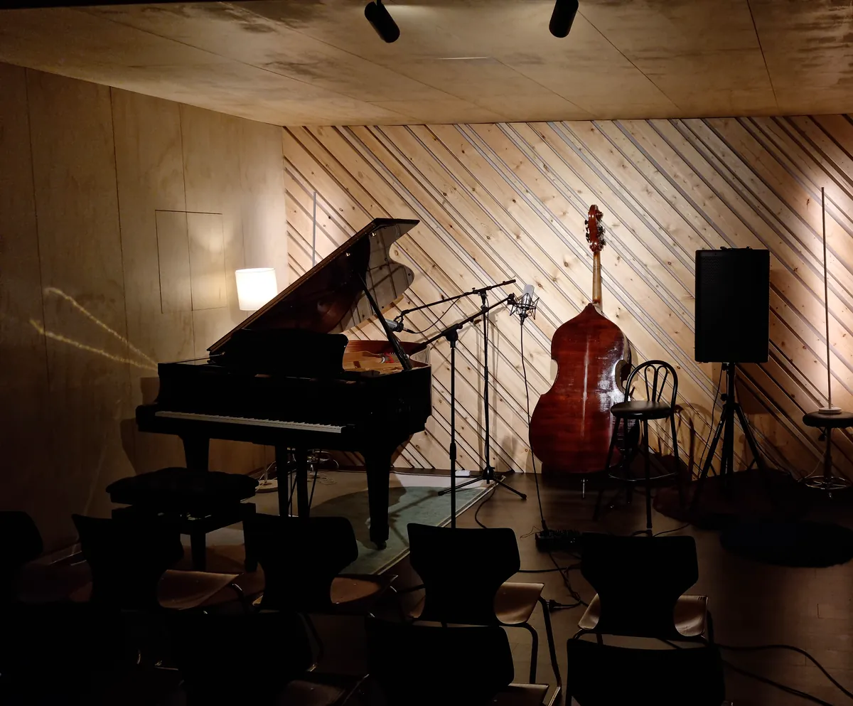 The hall stage in warm evening light: a grand piano, double bass, bar stool and speaker in front of the diagonal timber wall.
